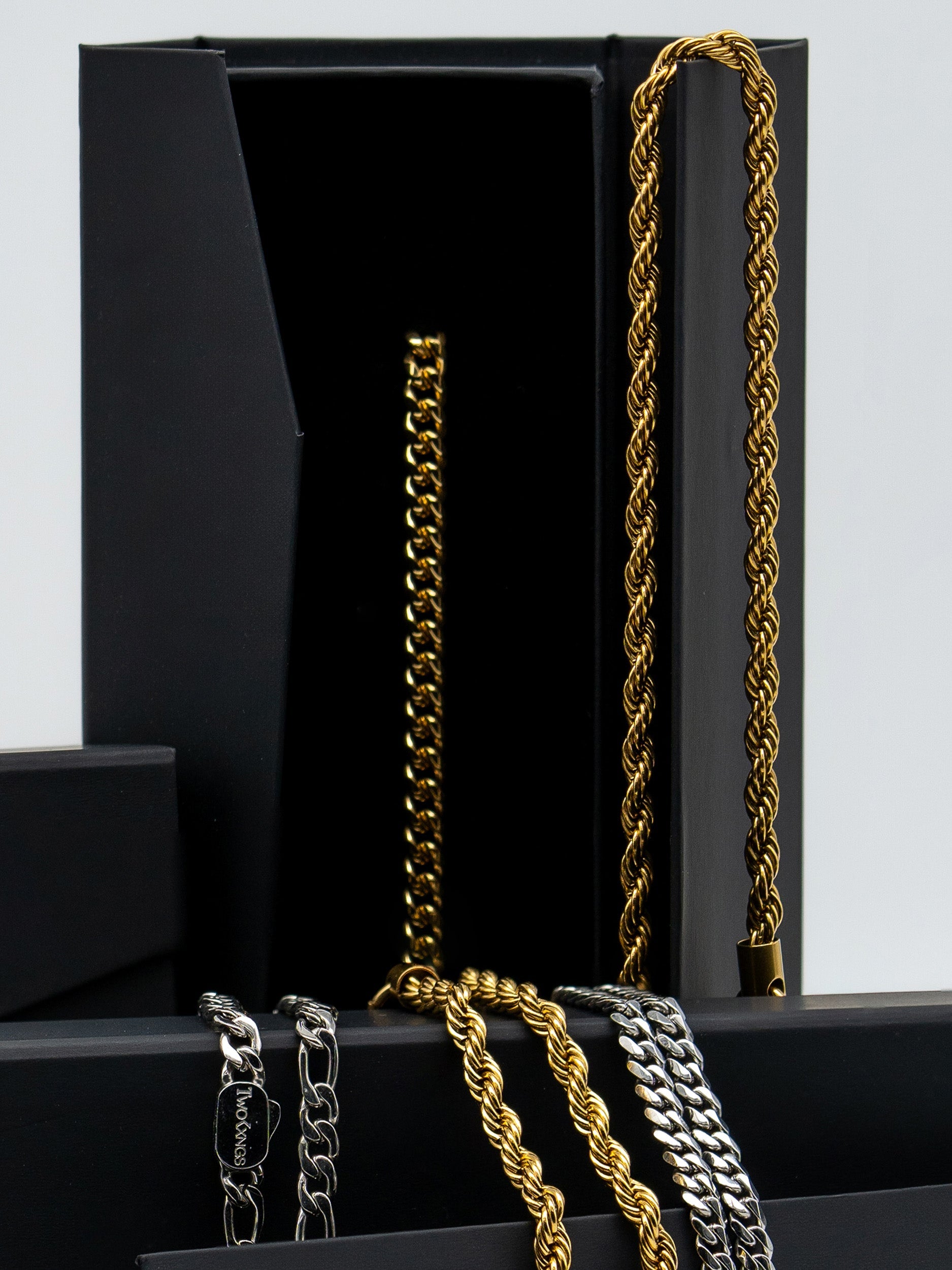 Two Kxngs chains in gold and silver draped over the black gift boxes, showing rope, cuban and figaro styles